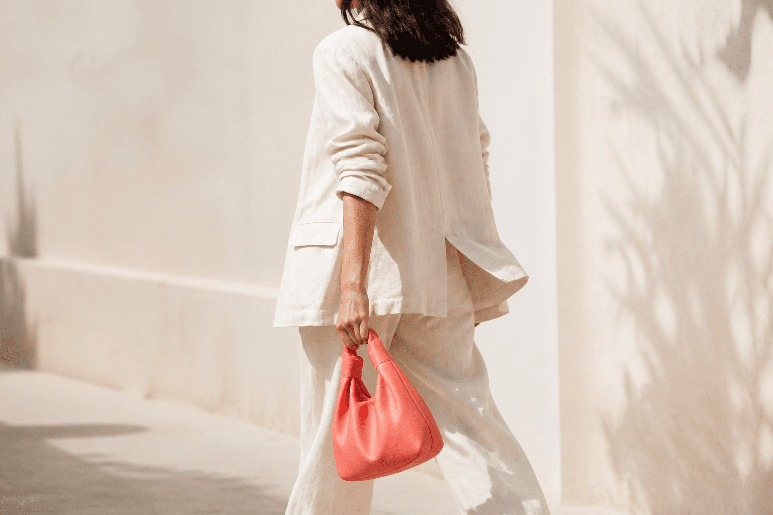 Person in a cream linen suit walking past a pale wall with a coral bag